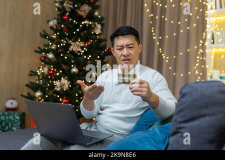 Verärgert weihnachtsmann mit Laptop versucht, Online-Kauf im Online-Shop zu machen, asiatische Mann mit Bankkarte erhielt Fehler und Betrug Nachricht, sitzt auf der Couch für das neue Jahr. Stockfoto