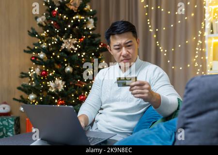 Verärgert weihnachtsmann mit Laptop versucht, Online-Kauf im Online-Shop zu machen, asiatische Mann mit Bankkarte erhielt Fehler und Betrug Nachricht, sitzt auf der Couch für das neue Jahr. Stockfoto