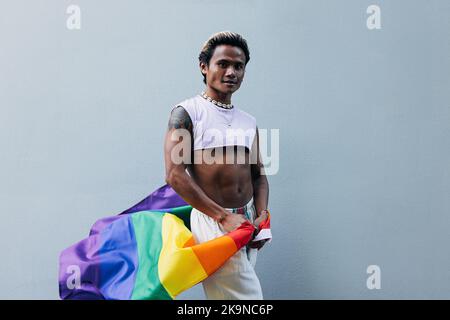 Junger Mann mit LGBT-Flagge, der draußen an einer grauen Wand posiert Stockfoto