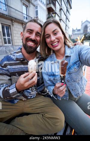 Foto eines glücklichen kaukasischen Paares, das in der Stadt Eis isst und ein Selfie mit Blick auf die Kamera macht. Stockfoto