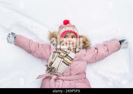 Kleines Mädchen in einer rosa Jacke mit einem überraschten Gesicht in einem Winterpark. Das Kind liegt in der Form eines Engels im Schnee Stockfoto