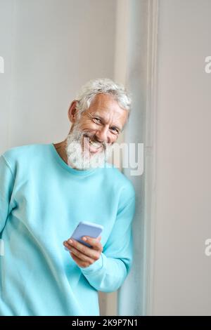 Glücklicher älterer bärtiger Hipster, der an einer weißen Wand mit einem Mobiltelefon stand. Stockfoto