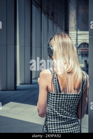 Rückansicht einer jungen blonden Frau, die über dem Ohr Kopfhörer trägt. Mädchen hört Musik auf der Straße Stockfoto