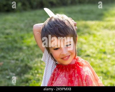 Porträt eines lächelnden Jungen, der American Indian spielt. Kind mit weißer Vogelfeder und rotem Umhang. Kostüm-Rollenspiel. Freizeitaktivitäten im Freien. Stockfoto