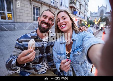 Foto eines glücklichen kaukasischen Paares, das in der Stadt Eis isst und ein Selfie mit Blick auf die Kamera macht. Stockfoto