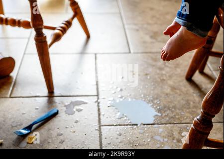 Kleinkind Fuß schwebte über verschüttete Milch, Krümel, und eine Kunststoffgabel auf einem braunen Fliesenboden Stockfoto