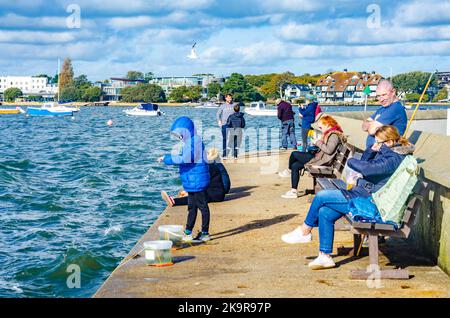 Eine Familie sitzt auf Bänken und beobachtet, wie ein Kind am Kai von Mudeford in der Nähe von Christchurch in Dorset, Großbritannien, Krabbenfischen genießt Stockfoto