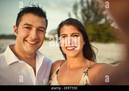 Selfies im Paradies. Beschnittenes Porträt eines liebevollen jungen Paares, das Selfies macht, während es am Strand steht. Stockfoto