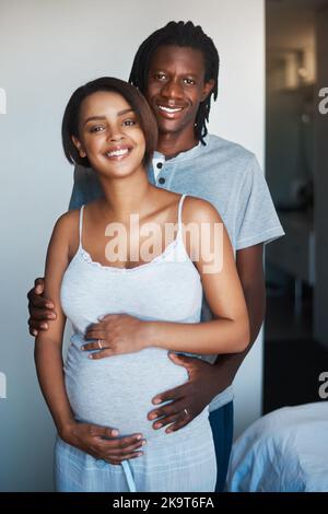 Das Lächeln auf unseren Gesichtern sagt alles. Porträt eines glücklichen jungen Mannes, der mit seiner schwanger Frau zu Hause posiert. Stockfoto