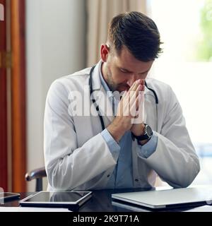 Betet für ein Wunder. Ein hübscher Arzt betet, während er in seinem Büro sitzt. Stockfoto