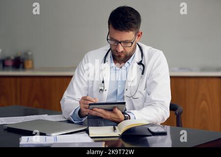 Mit Technologie, um Ihre Gesundheit zu verbessern. Ein hübscher männlicher Arzt, der auf seinem Tablet arbeitet, während er in seinem Büro sitzt. Stockfoto