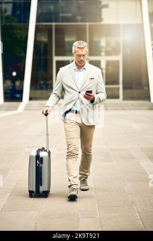 Das Geschäft hört nie auf, sich zu bewegen und auch nicht sein Ehrgeiz. Ein reifer Geschäftsmann, der ein Mobiltelefon benutzt, während er mit einem Koffer in der Stadt spazierengeht. Stockfoto