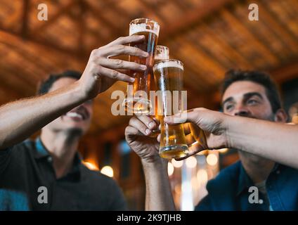 Diese geht zu uns hinaus. Drei fröhliche junge Männer, die während der Feier mit Bier an einem Tisch in einer Bierbrauerei sitzen Stockfoto