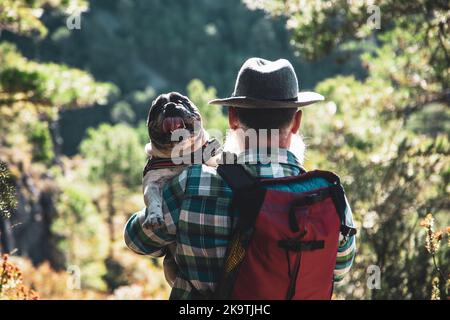 Ein hübscher bärtiger Mann, der lässig in der Natur herumläuft und seinen französischen Bulldoggen auf den Schultern trägt. Stockfoto