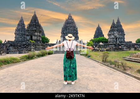 Indonesien, Prambanan – junges Mädchen mit Hut, der mit offenen Armen steht und während des Sonnenuntergangs den Prambanan-Tempel sieht. Asiatische Frau mit weißem Kleid und trägt ha Stockfoto