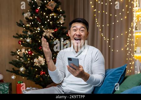 Weihnachtsporträt des erfolgreichen Mannes, des asiatischen Mannes, der die Kamera anschaut und die Hand hochhält, um Sieg und Triumphgesten zu gewinnen und auf dem Sofa in der Nähe des Weihnachtsbaums im Wohnzimmer zu Weihnachten und Neujahr zu sitzen. Stockfoto