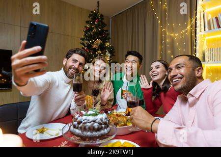 Diverse Freunde, die am Abend Weihnachten und Neujahr feiern, am Tisch sitzen, Wein trinken und essen, zusammen ein Selfie am Telefon in der Nähe des Weihnachtsbaums machen. Stockfoto