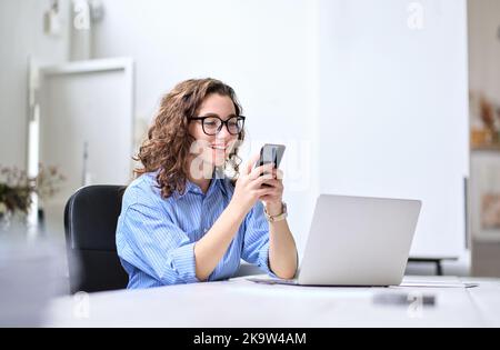 Glückliche junge Geschäftsfrau, die im Büro am Schreibtisch mit dem Handy arbeitet. Stockfoto
