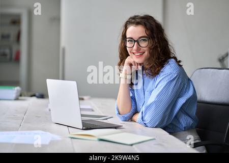 Junge glückliche Geschäftsfrau, die am Bürotisch sitzt und am Laptop arbeitet. Hochformat Stockfoto