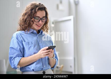 Lächelnde junge Geschäftsfrau mit Mobiltelefon, die im Büro mit dem Handy steht. Stockfoto