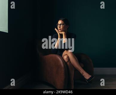 Schöne junge Frau schaut weg, während sie auf einem Stuhl sitzt. Junge asiatische Frau, die aus dem Fenster schaut, während sie in einer Bürolobby sitzt. Stockfoto