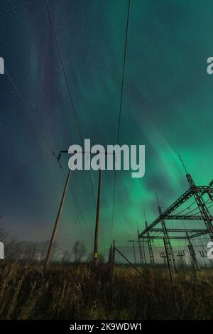 Schöne Nachtlichter über zwei hölzernen Pol-Stromleitungen und einem elektrischen Umspannwerk im Herbstfeld, Seitenansicht, vertikales Foto des Sternenhimmels mit Aur Stockfoto