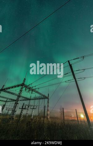 Wunderschöne Nachtlichter über einzelnen hölzernen Pol-Stromleitungen und einem elektrischen Umspannwerk im Herbstfeld, Seitenansicht, vertikales Foto des Sternenhimmels mit Aur Stockfoto