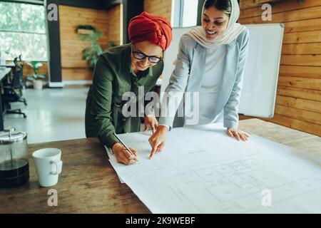 Fröhliche muslimische Architekten arbeiten in einem modernen Büro an Blaupause-Zeichnungen. Zwei kreative Geschäftsfrauen planen ein neues innovatives Projekt. Weibliche Desi Stockfoto