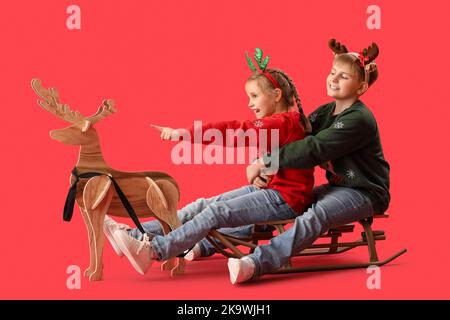 Glückliche kleine Kinder mit Schlitten und hölzernen Rentieren auf rotem Hintergrund Stockfoto