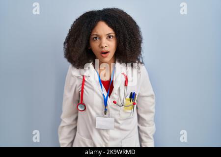 Junge afroamerikanische Frau in Arztuniform und Stethoskop im Schockgesicht, skeptisch und sarkastisch aussehend, überrascht mit offenem Mund Stockfoto