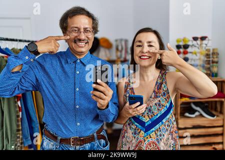 Interrassisches Paar im mittleren Alter im Einzelhandel mit Smartphone, das mit der Hand Finger auf Gesicht und Nase zeigt und fröhlich lächelt. Schönheitskonzept Stockfoto