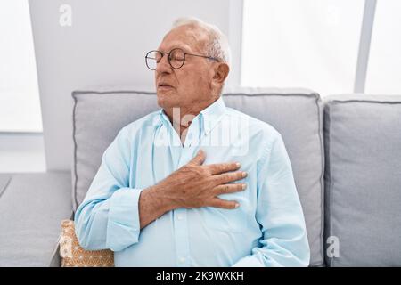 Älterer grauhaariger Mann, der an einem Herzinfarkt leidet und zu Hause auf dem Sofa sitzt Stockfoto