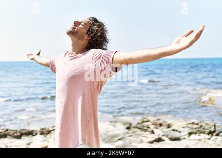 Junger hispanischer Mann atmet beim Stehen mit offenen Armen am Strand. Stockfoto