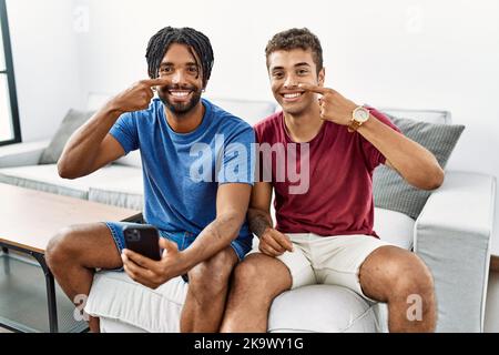 Junge hispanische Männer mit Smartphone sitzen auf dem Sofa zu Hause mit der Hand Finger zu Gesicht und Nase, lächelnd fröhlich. Beauty-Konzept Stockfoto