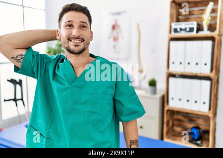 Der junge Physiotherapeut, der in der Schmerzklinik arbeitet, lächelt selbstbewusst und berührt die Haare mit der Hand nach oben, posiert attraktiv und modisch Stockfoto