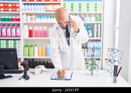 Junger kahlköpfiger Apotheker, der auf dem Smartphone spricht und auf einem Dokument in der Apotheke schreibt Stockfoto