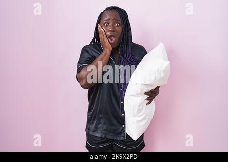 Junge afrikanische Frau trägt Pijama umarmt Kissen Angst und schockiert, Überraschung und erstaunt Ausdruck mit Händen auf dem Gesicht Stockfoto