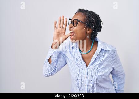 Afrikanische Frau mit Dreadlocks, die auf weißem Hintergrund stehen und eine Brille tragen, die laut auf der Seite mit der Hand auf dem Mund schreit und schreit Stockfoto