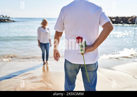 Mann und Frau im mittleren Alter überraschen am Meer mit Blumen auf dem Rücken Stockfoto