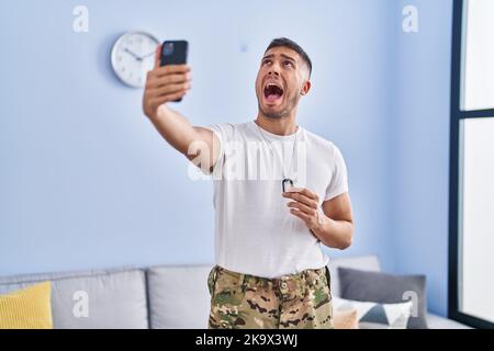 Junger hispanischer Mann in Tarnuniform nimmt Selfie zu Hause auf wütend und wütend schreiend frustriert und wütend, schreiend mit Wut und Blick U Stockfoto