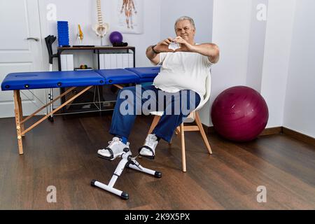 Leitender kaukasischer Mann in der Physiotherapie-Klinik mit Pedal-Übungsgerät lächelnd in der Liebe dabei Herz Symbol Form mit Händen. Romantisches Konzept. Stockfoto