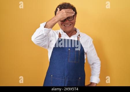 hispanischer Mann mittleren Alters mit professioneller Kochschürze, lächelnd und lachend, mit der Hand auf dem Gesicht, die die Augen bedeckt, um zu überraschen. Blind-Konzept. Stockfoto