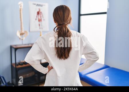 Junge Brünette Frau, die in der Klinik für Schmerzwiederherstellung arbeitet, steht rückwärts und blickt weg mit den Armen am Körper Stockfoto