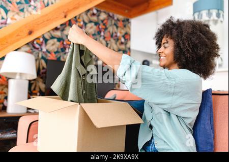 Positive junge schwarze Frau, die mit einem Karton steht und neue Kleidung auszieht, während sie neugierig auf bestellte Artikel aus dem Online-Shop ist. Lächelnder Kunde zufrieden mit schnellem Kurierdienst Stockfoto
