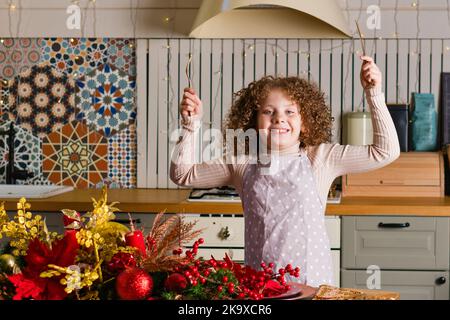 Fröhliche lockige Mädchen in Schürze steht in der Küche am Tisch für Weihnachten geschmückt und hält Besteck. Kind hält Gabel und Messer. Bereit für das Feiertagsessen. Stockfoto