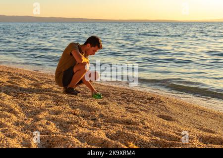 Tag der Erde. Müllabfuhr an der Adriaküste. Ein Junge, der am Strand eine Plastikflasche abholt. Sonnenuntergang. Das Konzept der Erhaltung der Ökologie Stockfoto