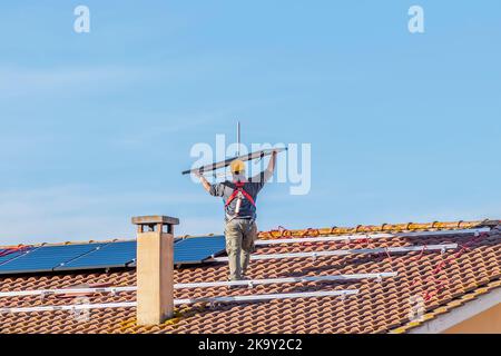 Ein Techniker hebt ein Solarpanel über seinen Kopf, während er sich darauf vorbereitet, es auf dem Dach eines rot gefliesten Hauses zu montieren Stockfoto