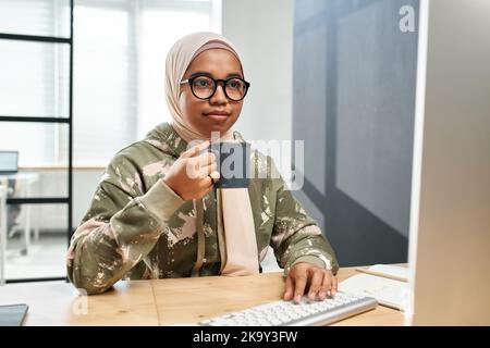 Junge seriöse IT-Ingenieurin in Hijab, Casualwear und Brillen, die Kaffee vor dem Computermonitor an ihrem Arbeitsplatz im Büro trinken Stockfoto