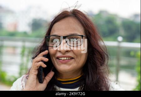 Lächelnde Frau mit langen braunen Haaren bei mittleren Erwachsenen, die im Freien auf dem Mobiltelefon spricht. Nahaufnahme. Sie trägt weißes T-Shirt und Specs. Vorderansicht. Kopf und Stockfoto