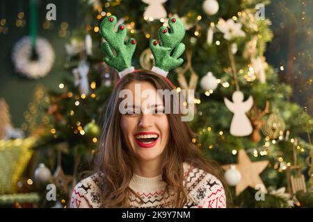 Weihnachtszeit. Porträt einer glücklichen Frau mittleren Alters mit lustigen Rentier Geweih Kopfschmuck in traditionellen Weihnachts-Pullover in der Nähe von Weihnachtsbaum in Stockfoto